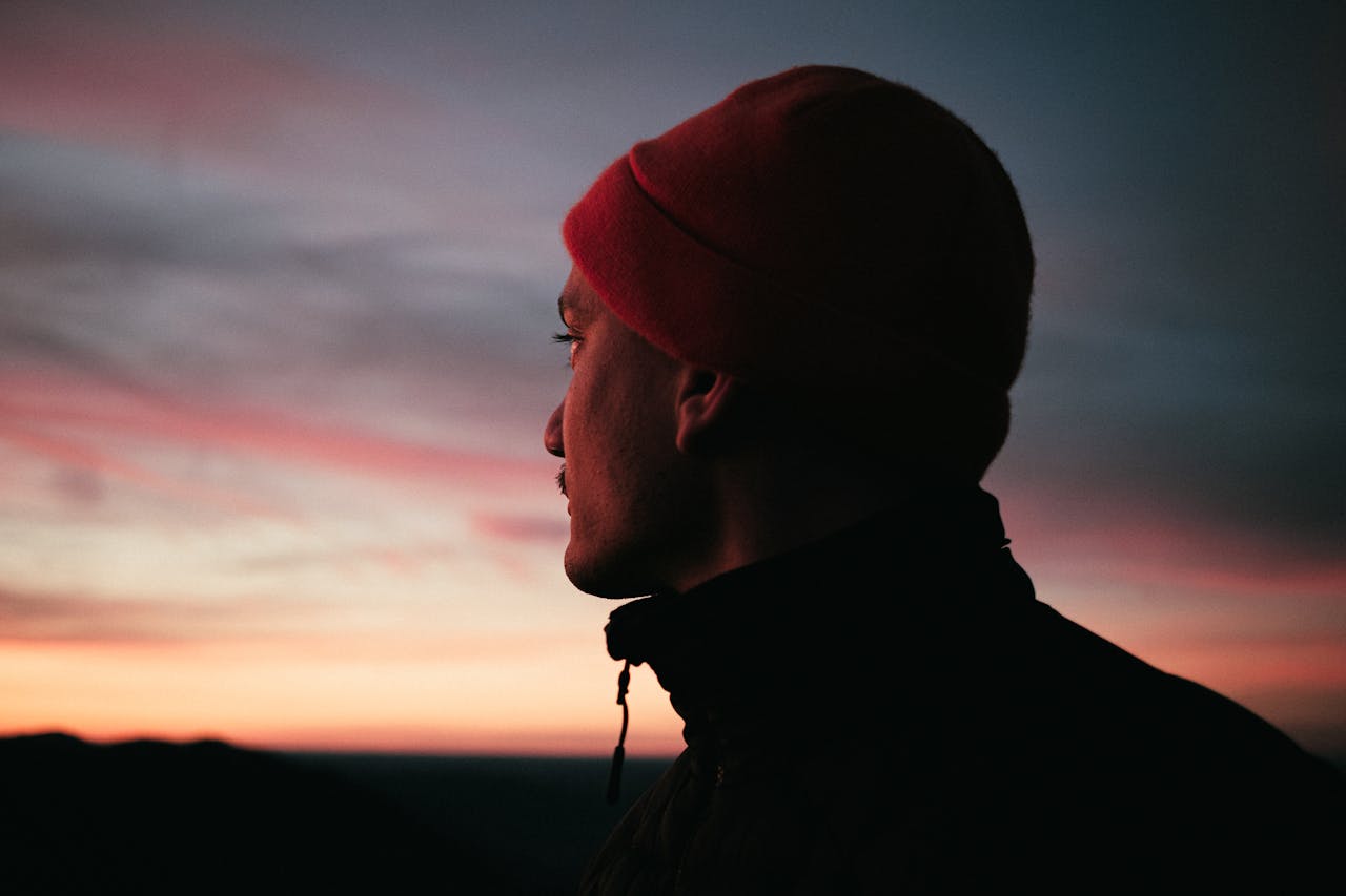 A man in a beanie looks away, silhouetted against a colorful sunset in Deutschland.